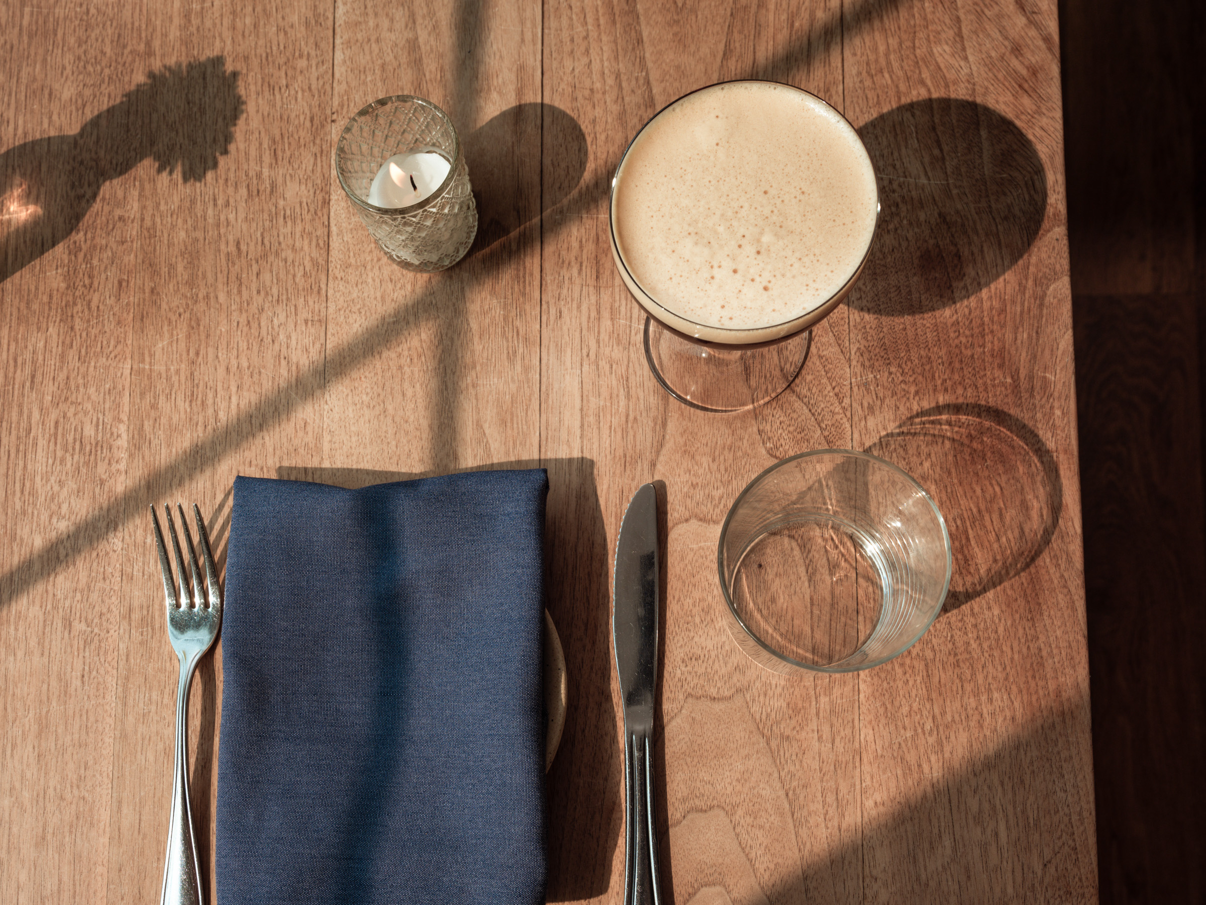 A top-down view of a dining table setting featuring a neatly folded navy napkin, a fork and knife, a glass of cocktail, and a candle in a decorative holder, with soft shadows cast by natural light.