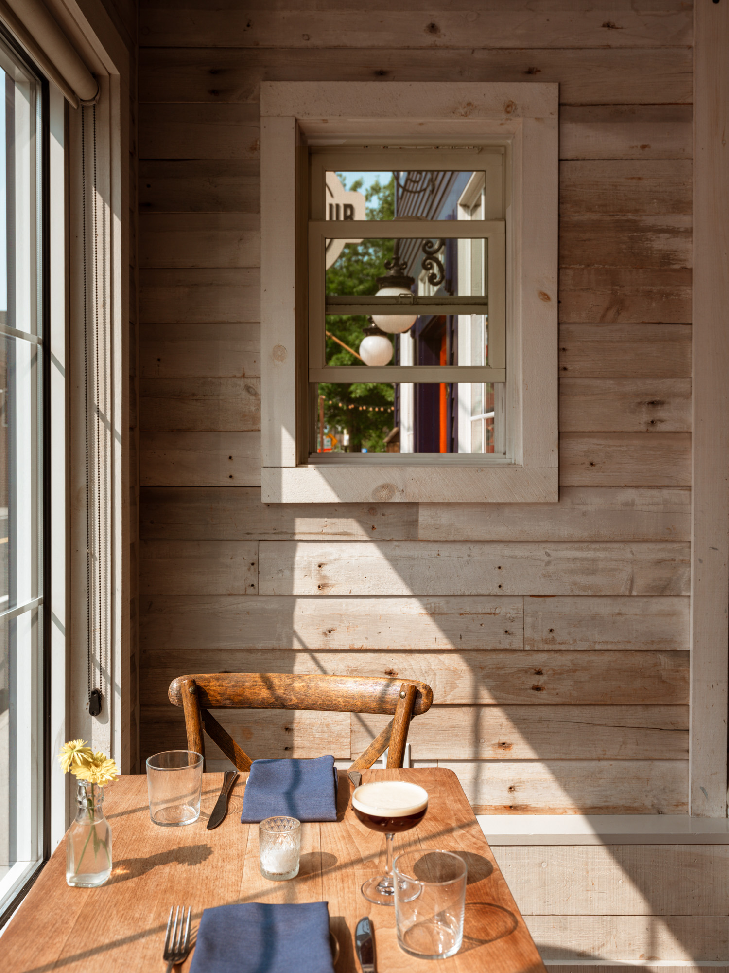 A cozy dining setting featuring a wooden table with blue napkins, glasses, and a flower vase, illuminated by natural light through a window.