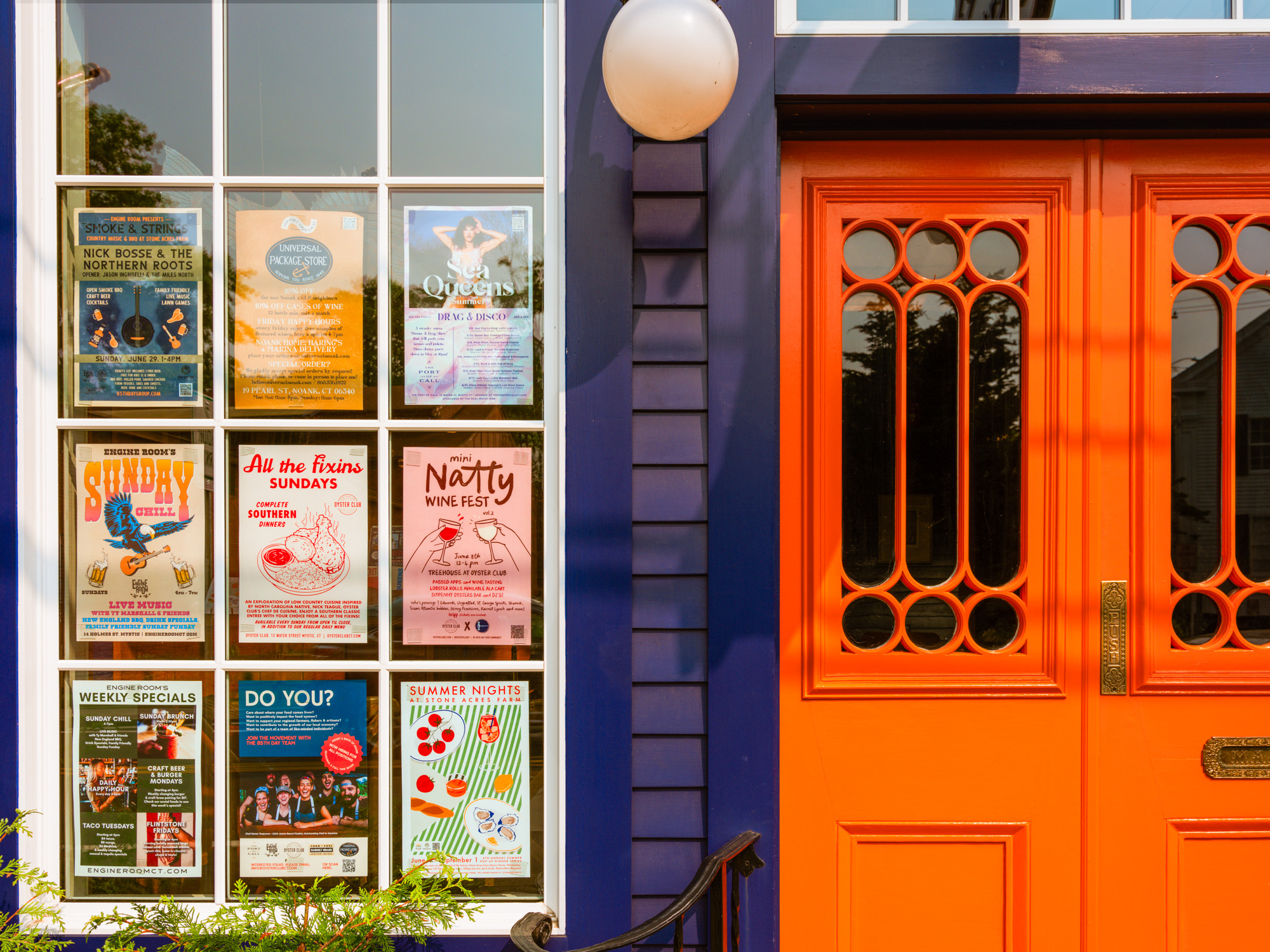 A vibrant orange door with decorative glass panels, framed by a window displaying various colorful event posters, set against a blue building.