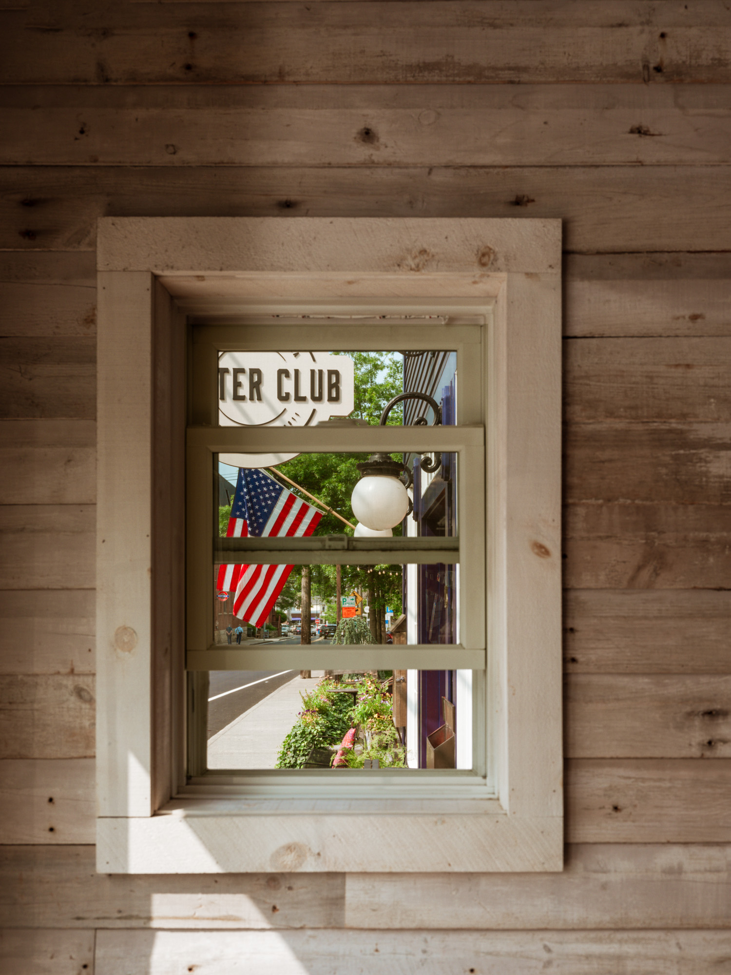 A view through a wooden-framed window showcasing an American flag and a vibrant street scene with greenery and a sign for a club.