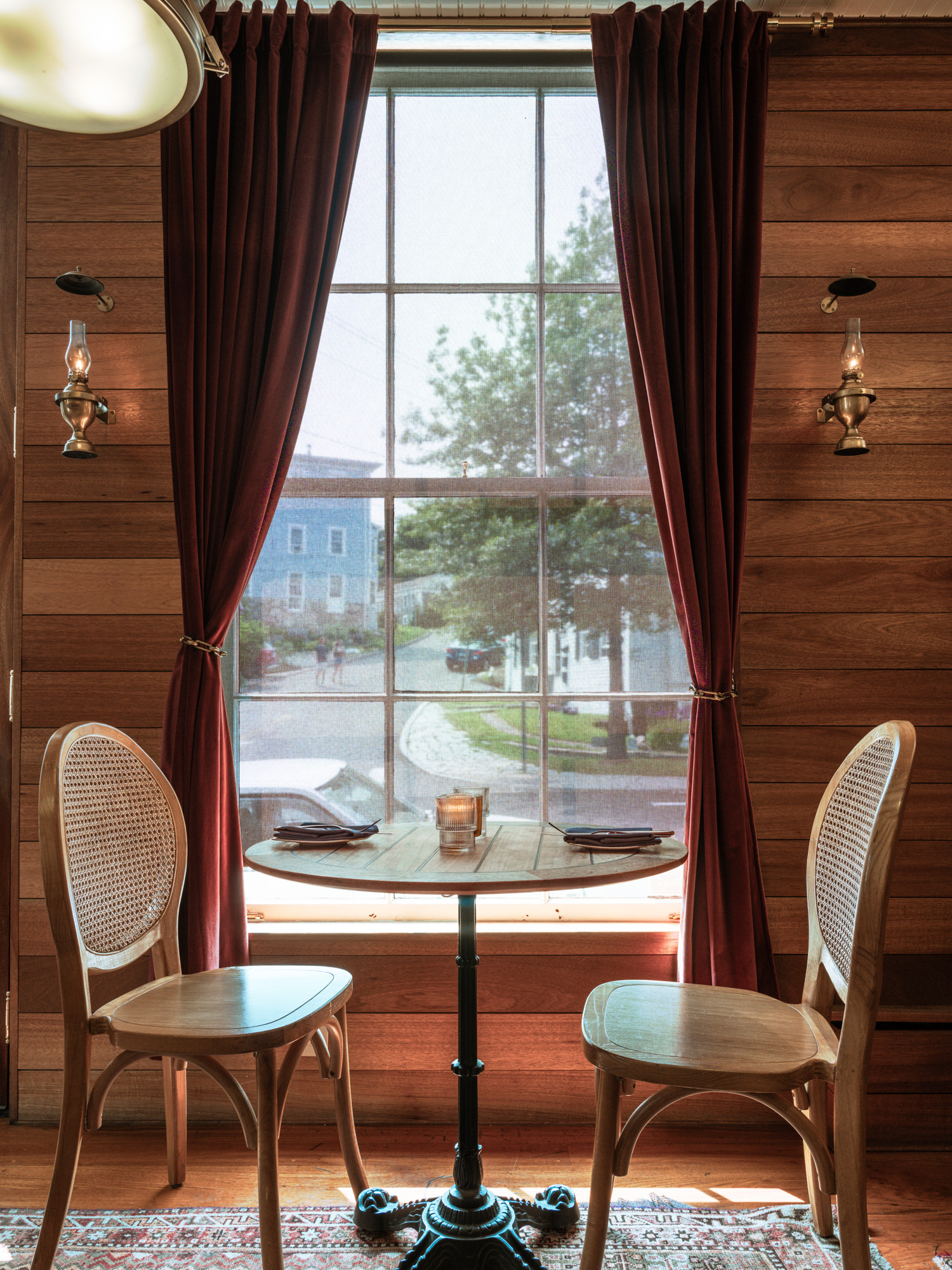 A cozy dining setup featuring two wooden chairs and a round table by a large window with red curtains, showcasing a serene outdoor view.