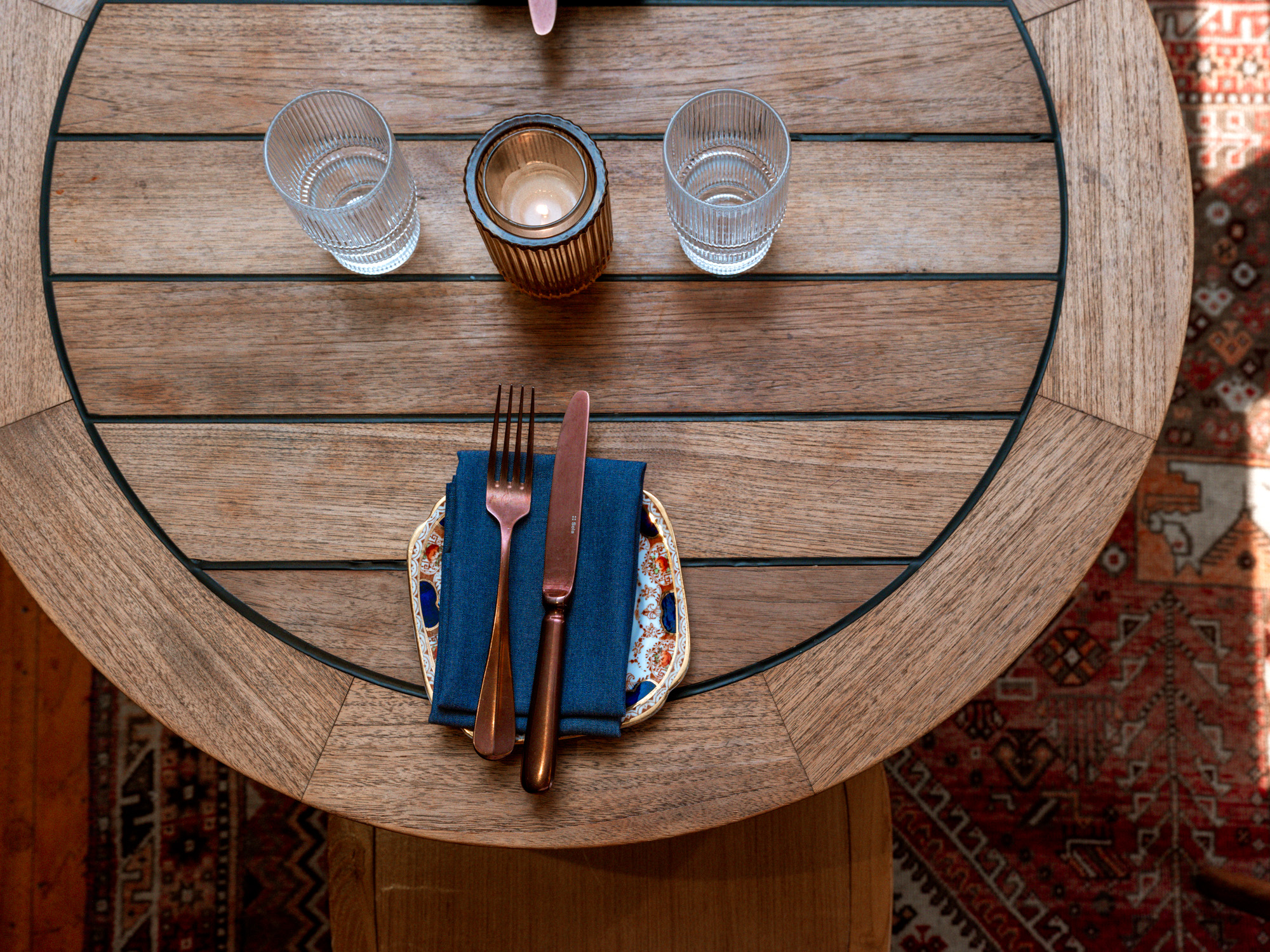 A beautifully set dining table featuring a round wooden table with a patterned tablecloth, two glasses of water, a candle, and neatly arranged cutlery and napkin.