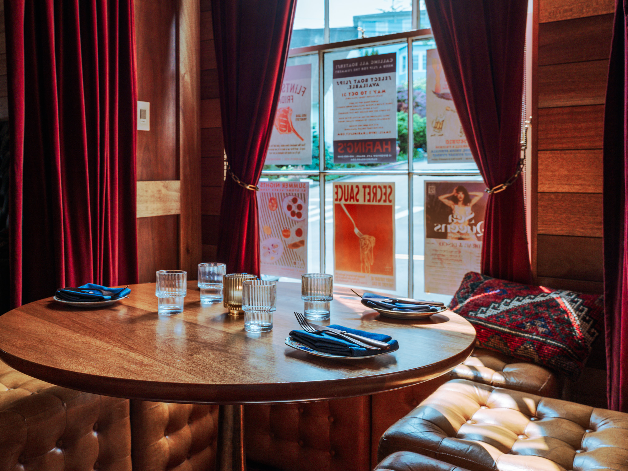 A cozy restaurant interior featuring a wooden table set for dining with blue napkins, surrounded by plush seating and rich red curtains. Posters are visible through the window.