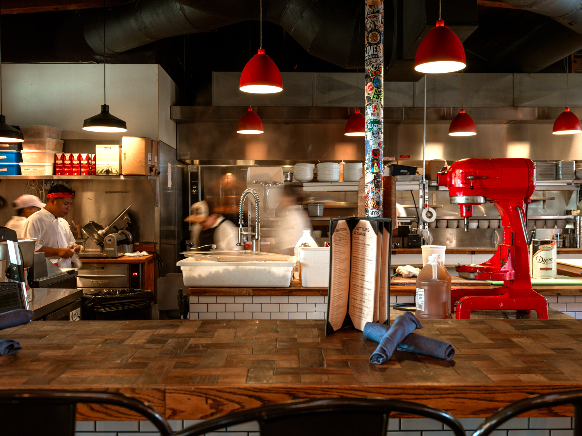 A bustling restaurant kitchen with chefs at work, featuring a red stand mixer and a wooden countertop. The setting is warm and inviting with pendant lighting.