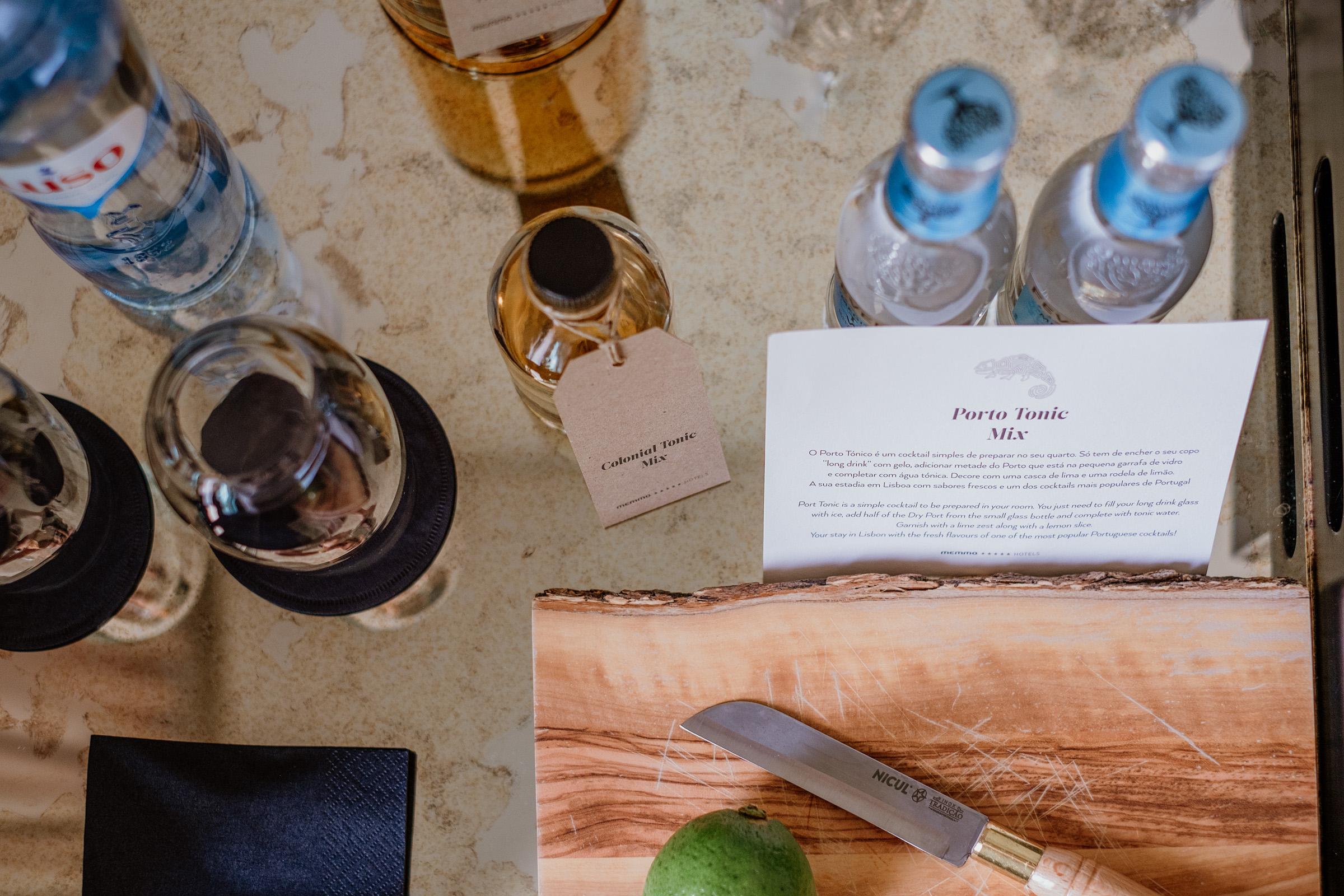 A top-down view of a wooden cutting board with a knife, a lime, and various bottles of spirits and mixers, including a recipe card for a Porto Tonic mix.