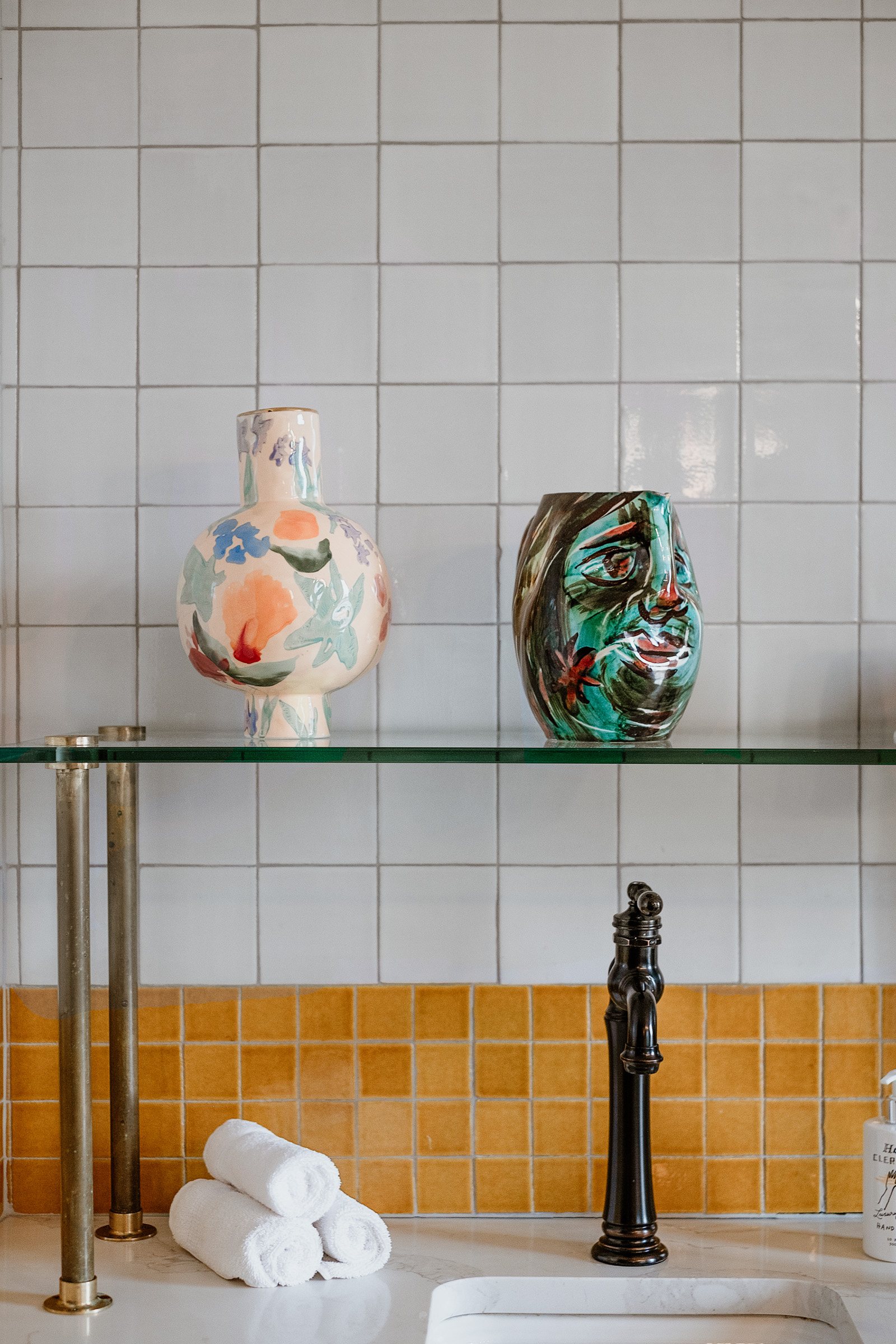 A stylish display of two decorative vases on a glass shelf against a tiled wall. One vase features floral patterns, while the other has a colorful face design. Below, neatly rolled white towels are placed beside a black faucet.
