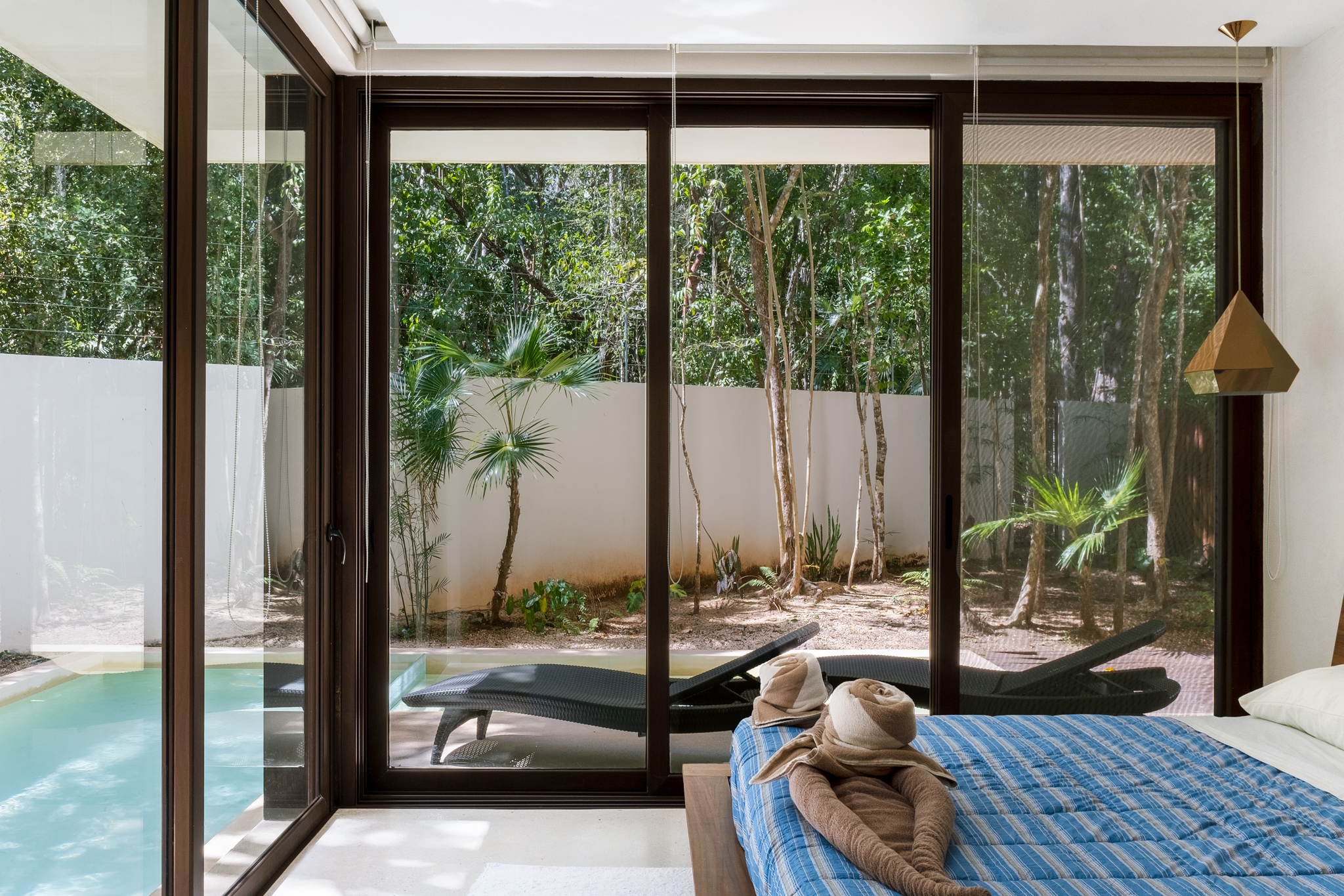 A modern bedroom with large glass windows overlooking a lush garden and pool area. The room features a neatly made bed with blue linens and decorative towel art.