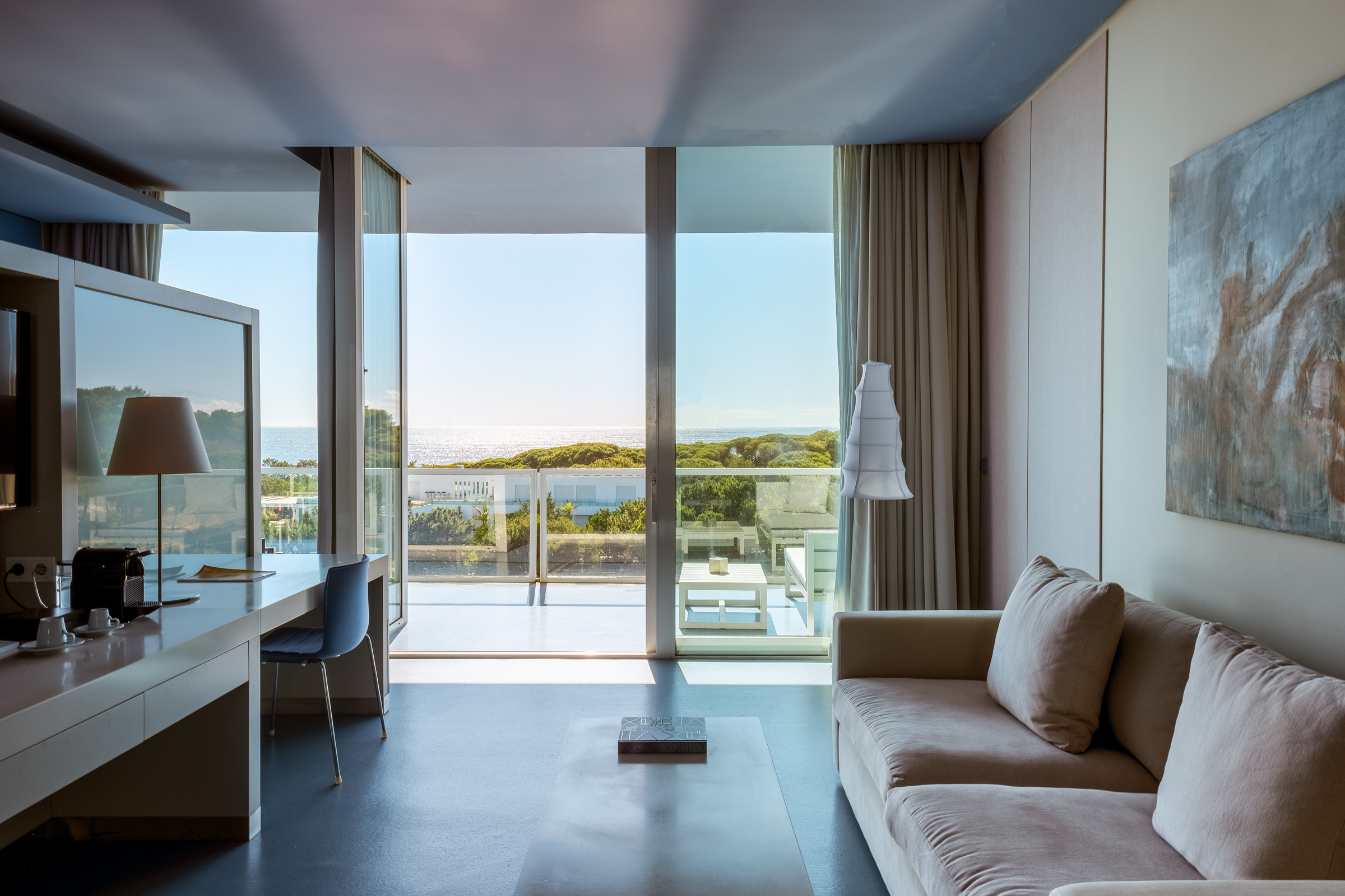 A modern hotel room featuring a comfortable beige sofa, a sleek desk, and large glass windows offering a view of the ocean and greenery outside.