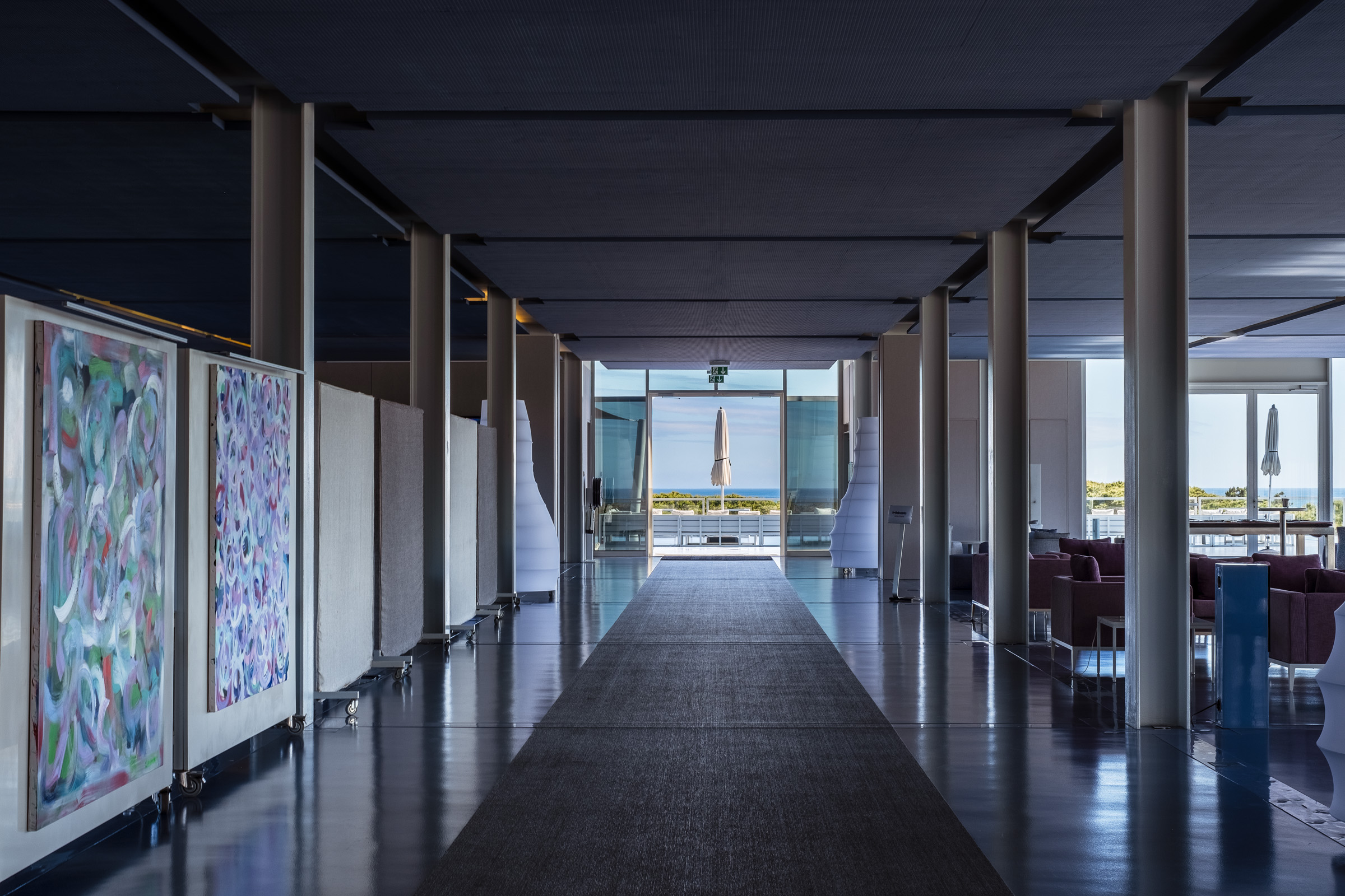A modern hotel corridor featuring abstract art on walls, leading to a large glass window with a view of the outdoors.