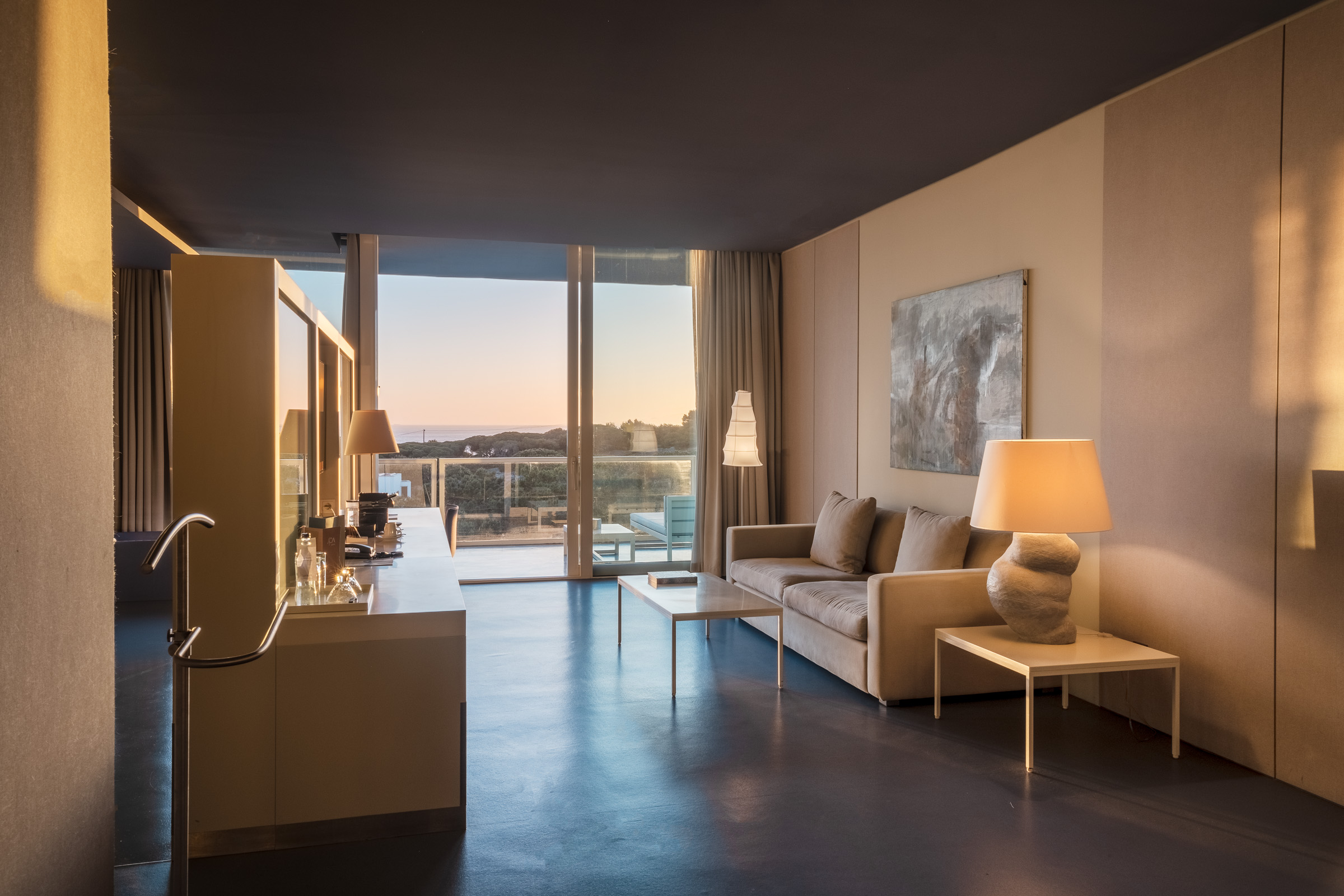 A modern hotel room featuring a beige sofa, a glass coffee table, and a lamp, with large windows showcasing a sunset view.