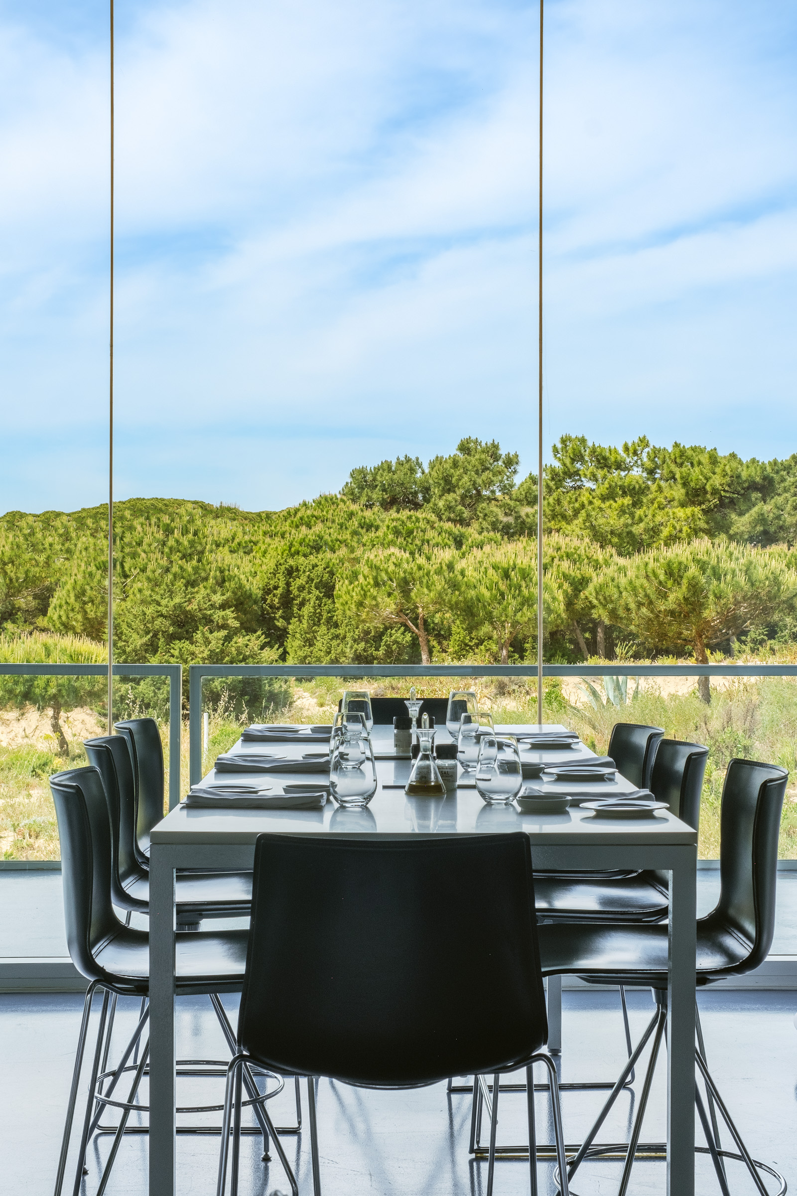 A modern dining table set for a meal, featuring black chairs and glassware, with a backdrop of lush greenery and a clear blue sky.