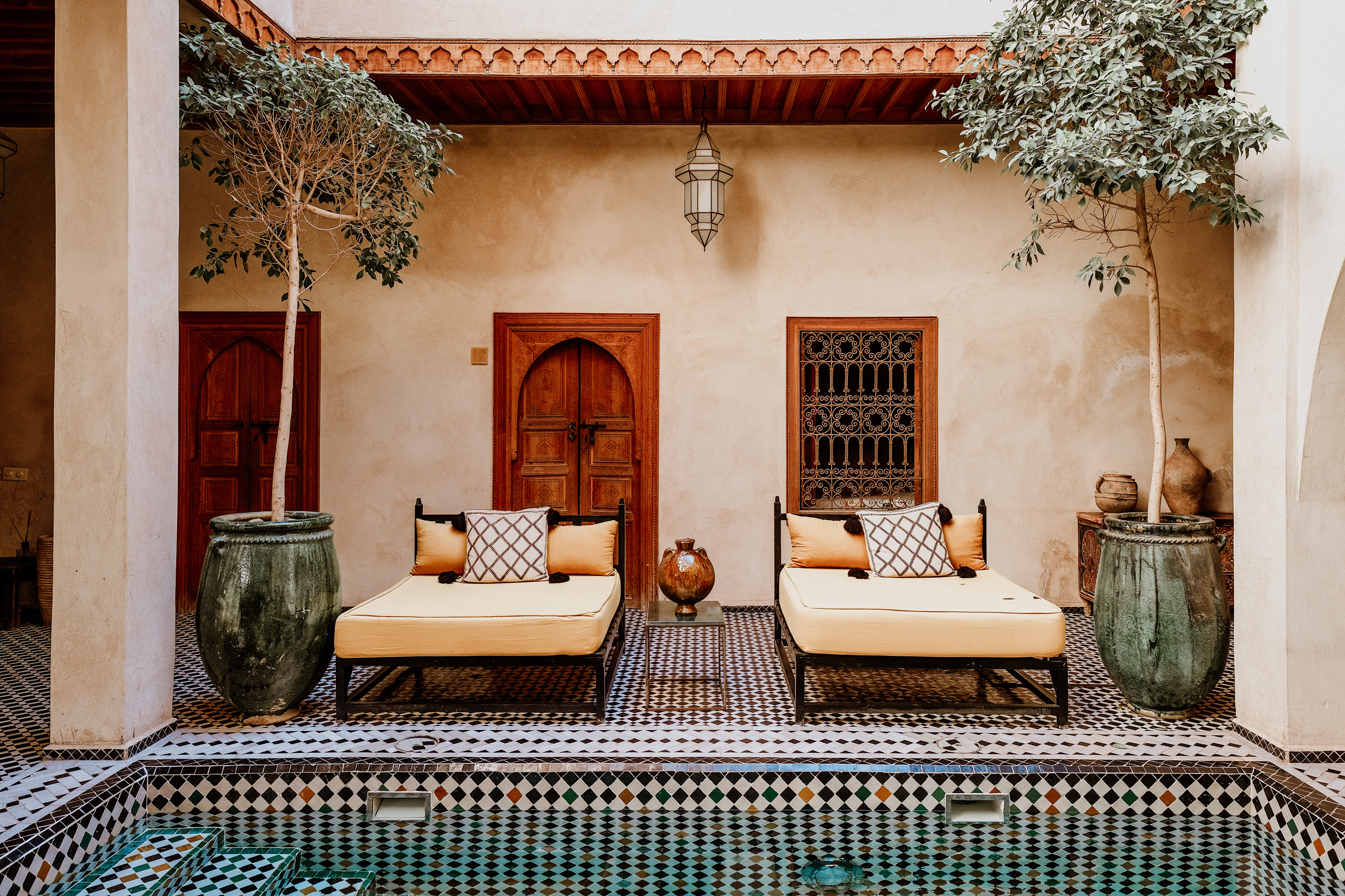 A serene courtyard featuring two yellow lounge chairs, surrounded by potted trees and intricate tile work, with a small pool reflecting the warm tones of the setting.