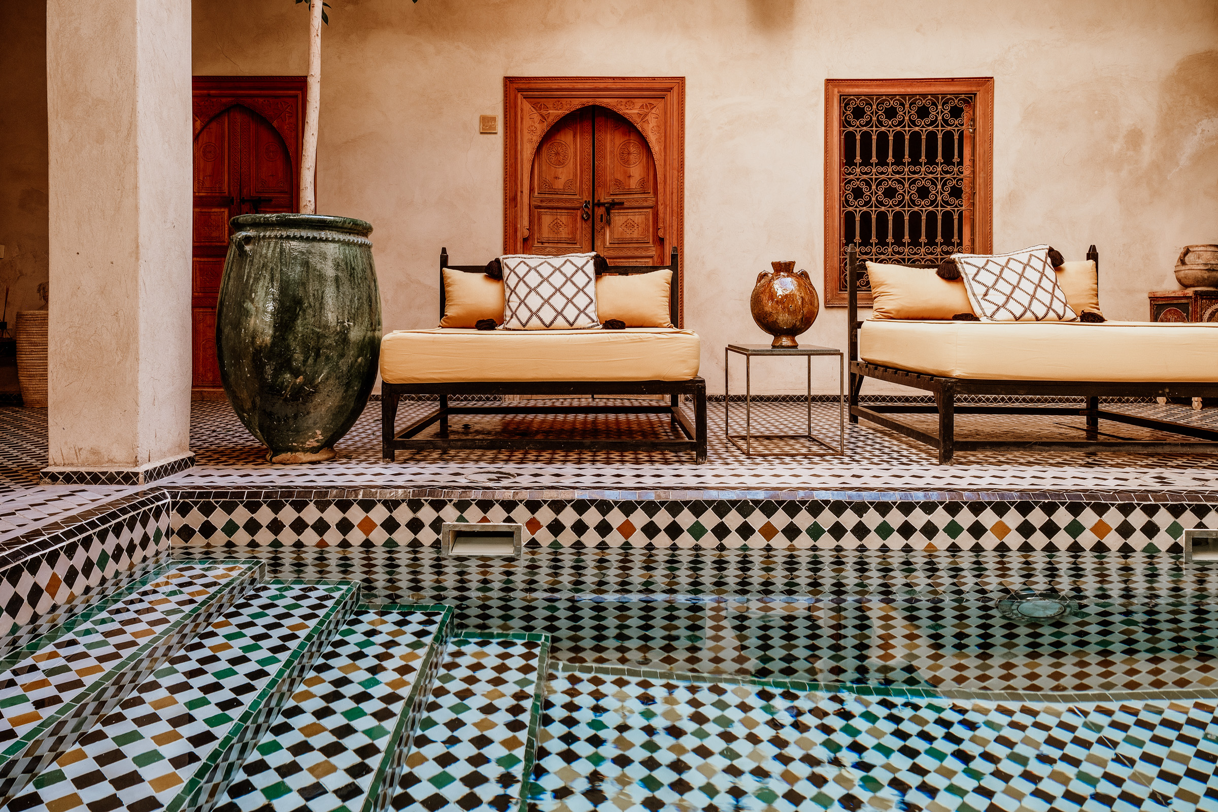 A serene courtyard featuring two elegant lounge chairs with cushions, a decorative pot, and a tiled pool with patterned steps, set in a Moroccan-style architecture.