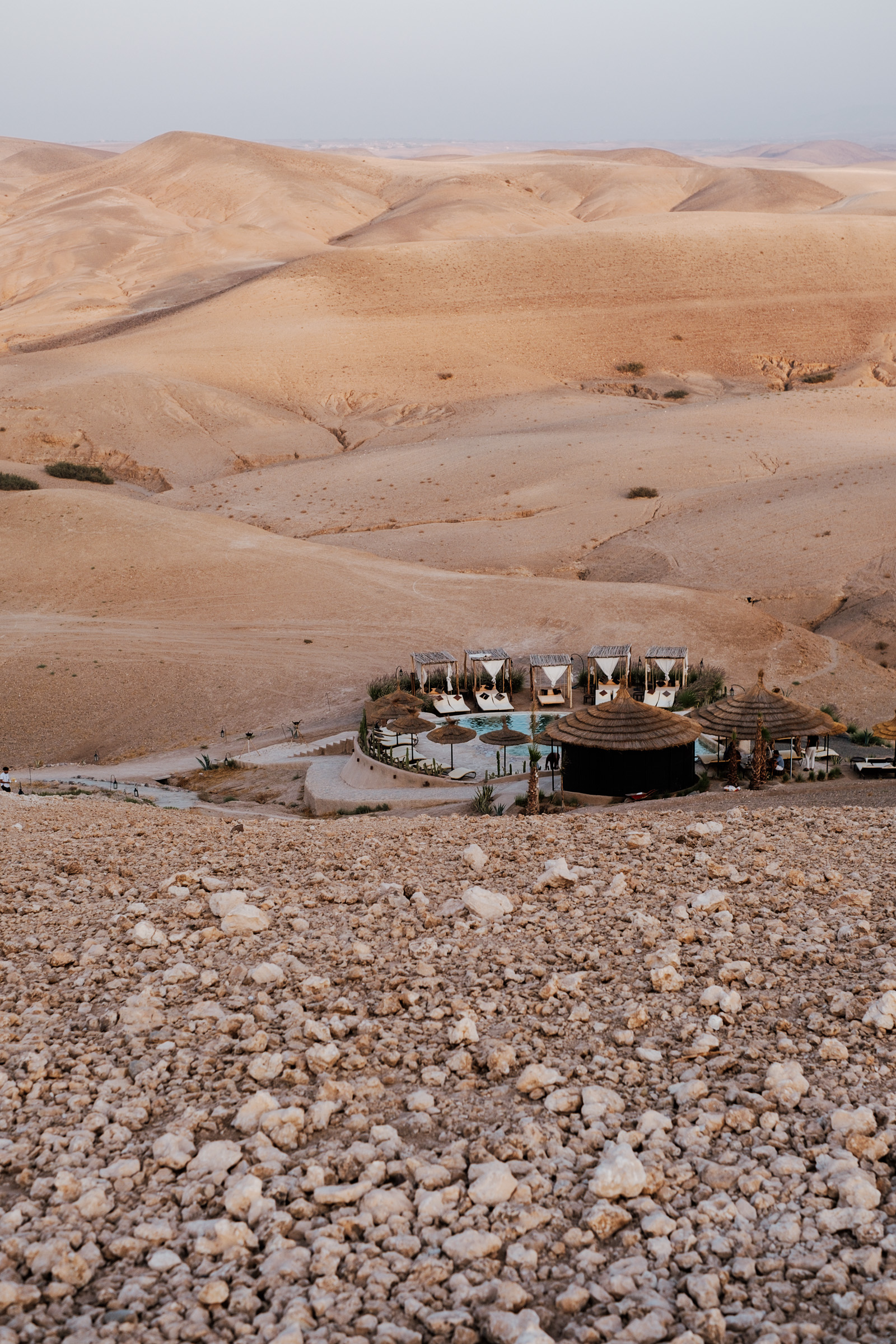 A panoramic view of a desert landscape featuring a resort with a pool, surrounded by arid hills and rocky terrain under a soft, warm light.