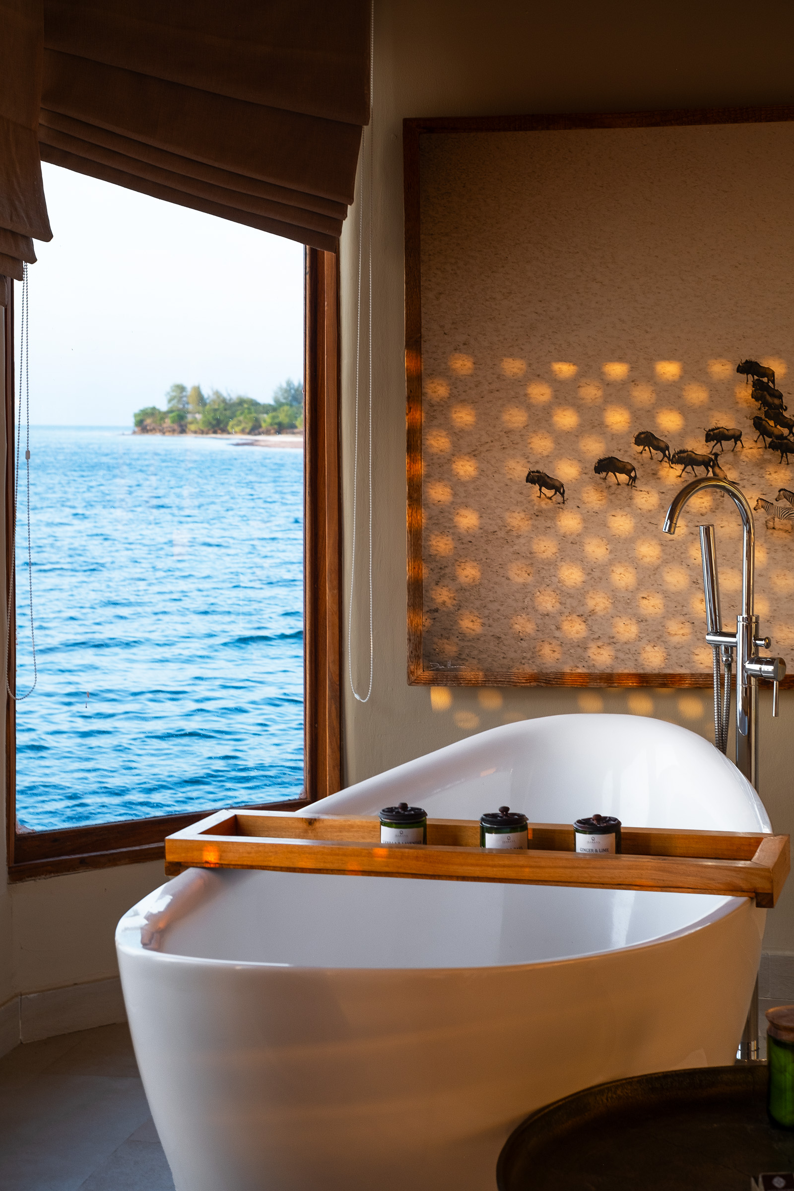 A serene bathroom with a freestanding white bathtub beside a large window overlooking a tranquil body of water. The setting features natural light casting patterns on the wall, enhancing the peaceful atmosphere.