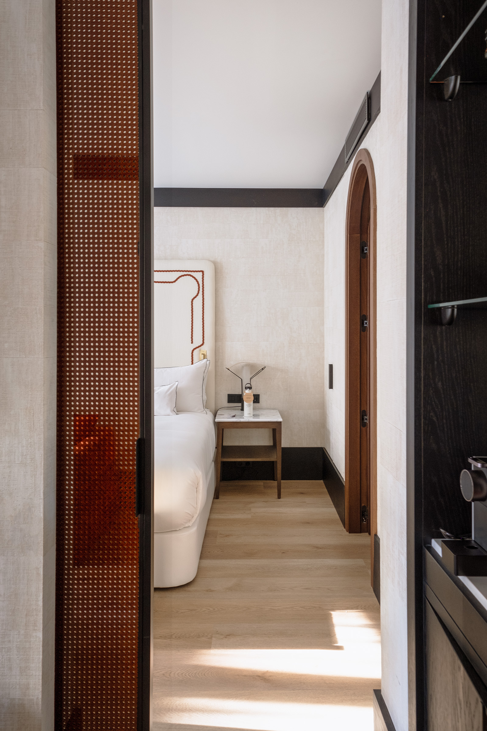 A modern hotel room featuring a minimalist design with a wooden floor, a white bed, and a small bedside table. The room is well-lit with natural light and has a warm, inviting atmosphere.