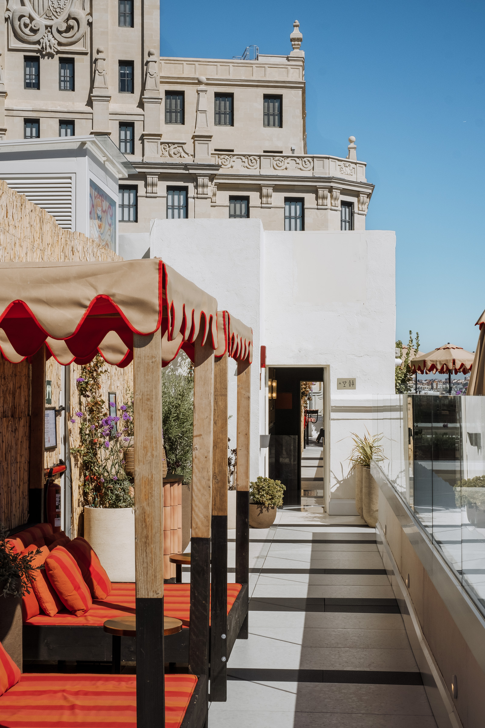 A stylish outdoor lounge area with red and beige canopies, featuring comfortable seating and a view of a historic building under a clear blue sky.