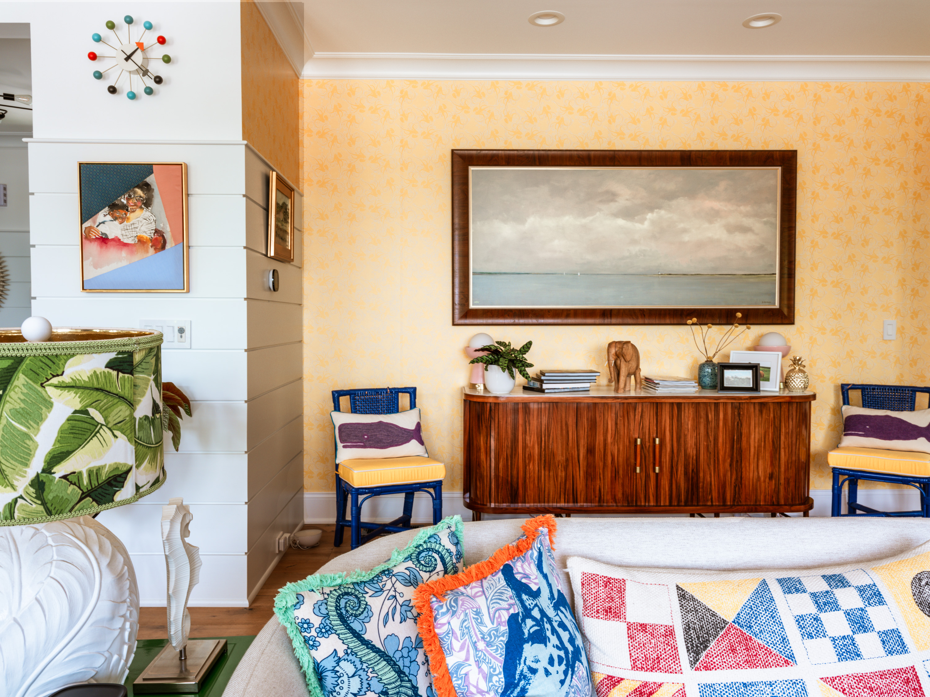 A cozy living room featuring a wooden sideboard with a landscape painting above it, colorful chairs, and decorative pillows on a sofa.