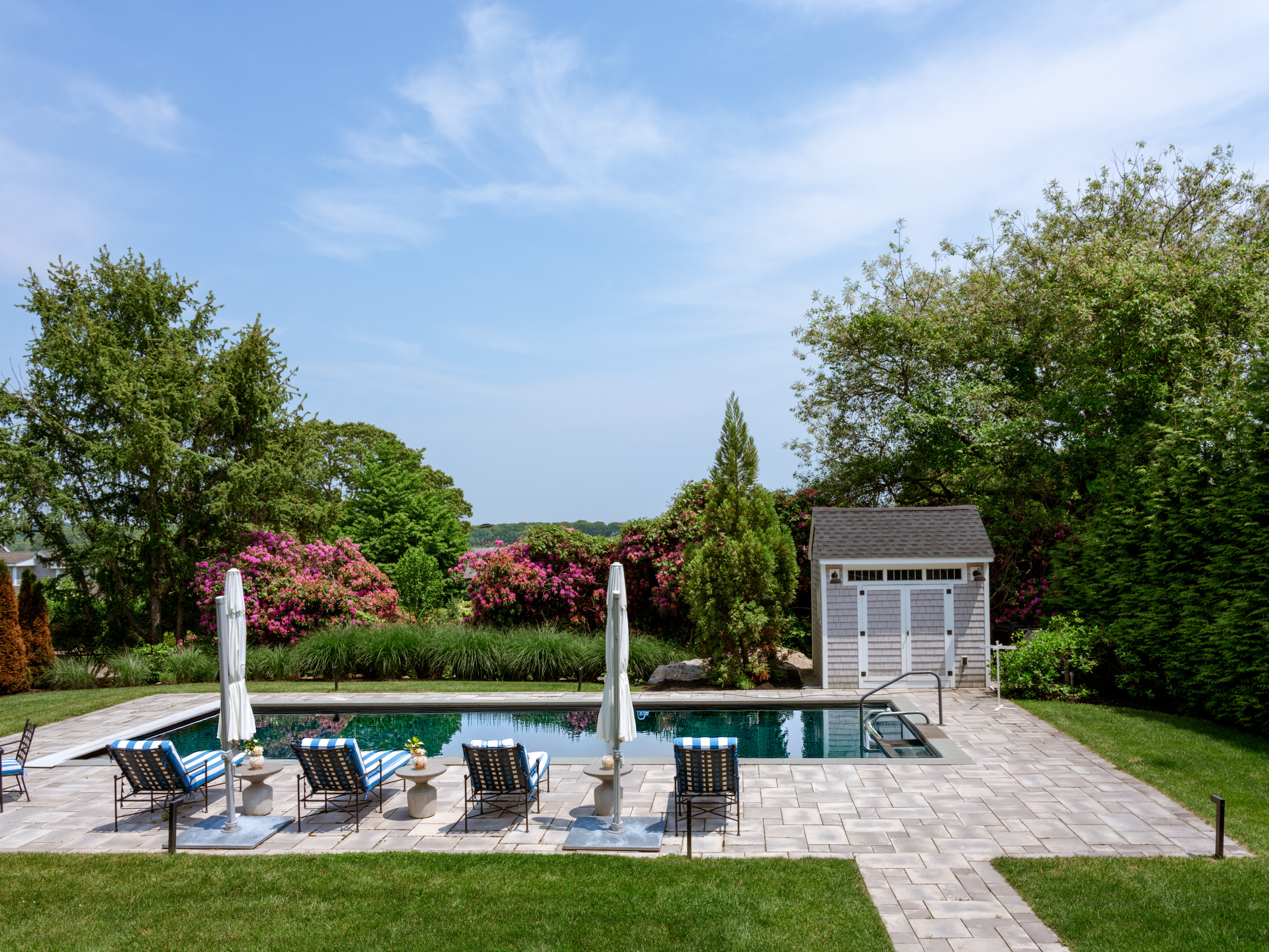 A serene backyard pool area with lounge chairs, surrounded by lush greenery and blooming flowers, under a clear blue sky.
