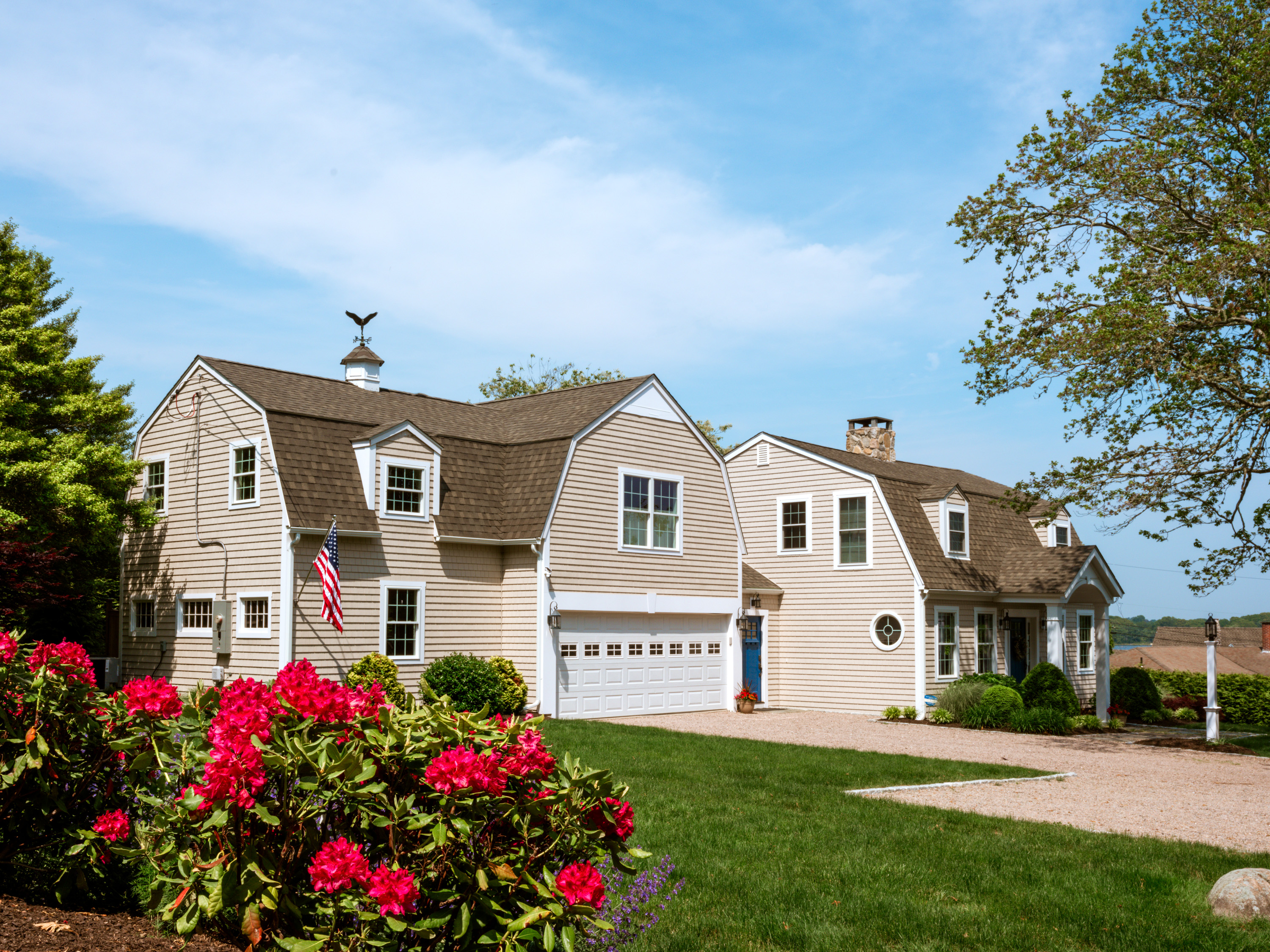 A charming beige house with a well-manicured lawn, vibrant pink flowers in the foreground, and a clear blue sky in the background.
