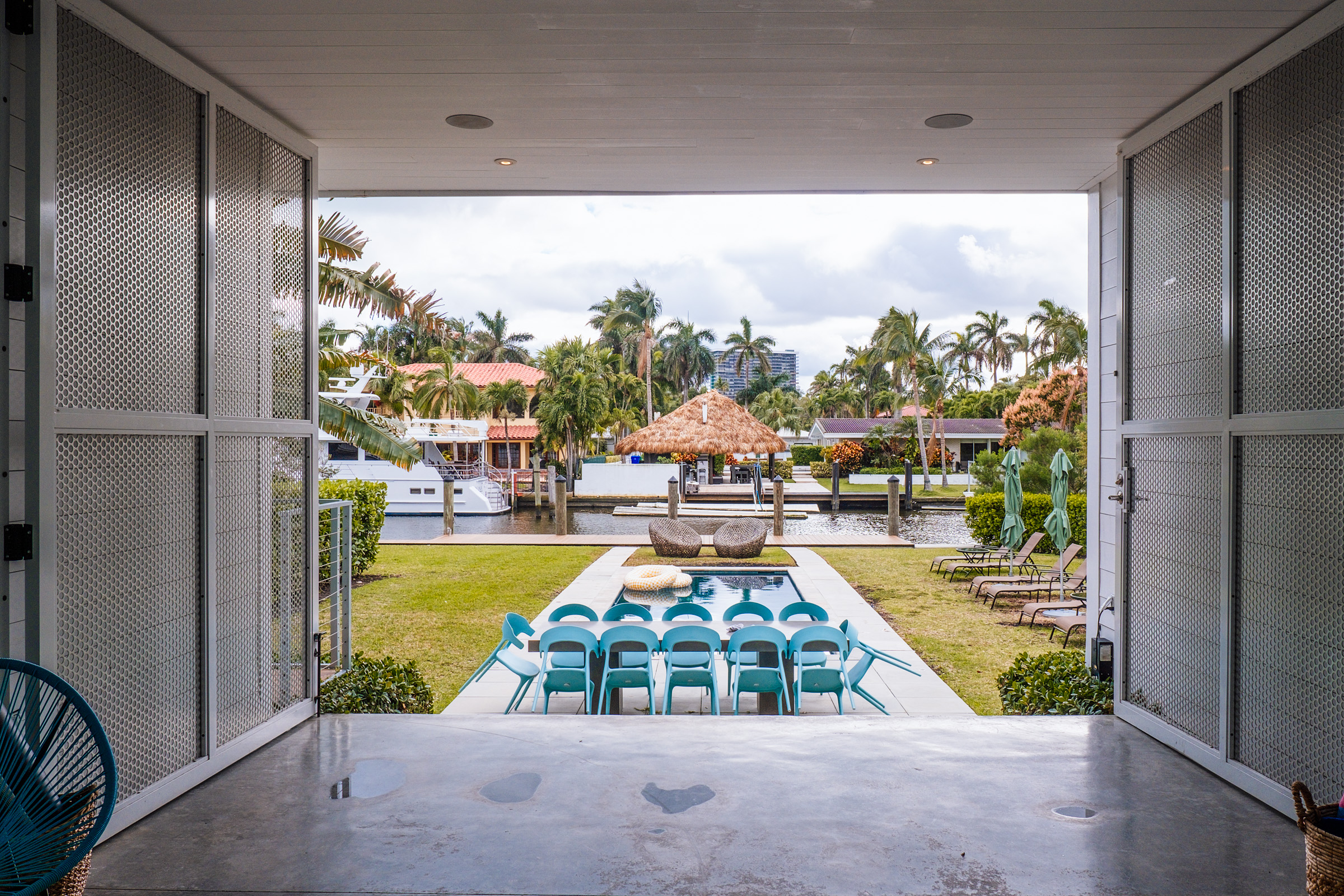 A view from an open doorway showcasing a pool area with blue chairs, a thatched gazebo, and palm trees in a tropical setting.