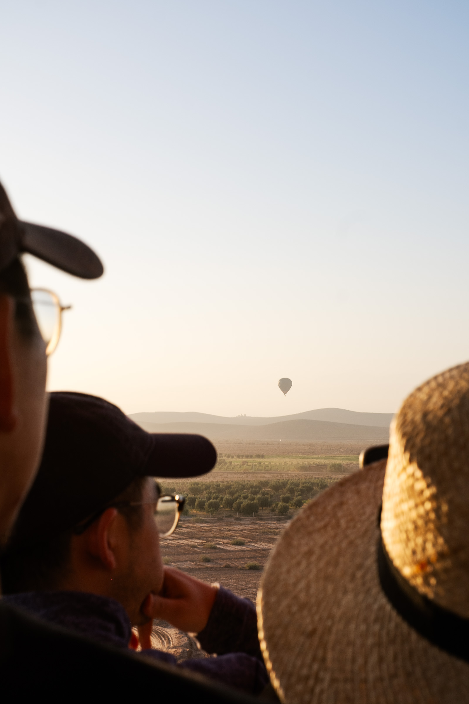 A person wearing glasses and a straw hat watches a hot air balloon in the distance during sunrise, with rolling hills and fields in the background.