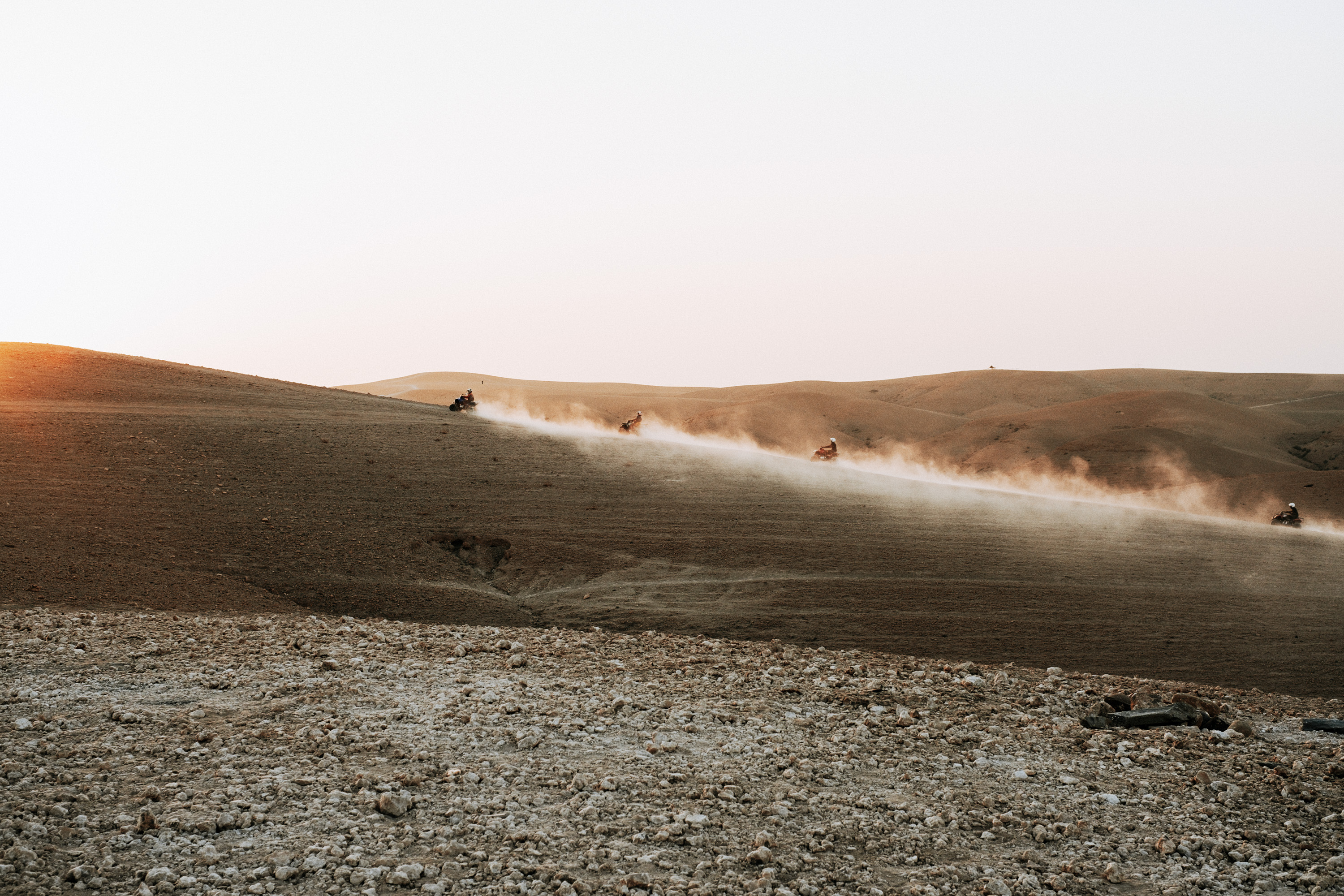 A group of off-road vehicles kicking up dust while traversing a sandy landscape at sunset, with rolling hills in the background.