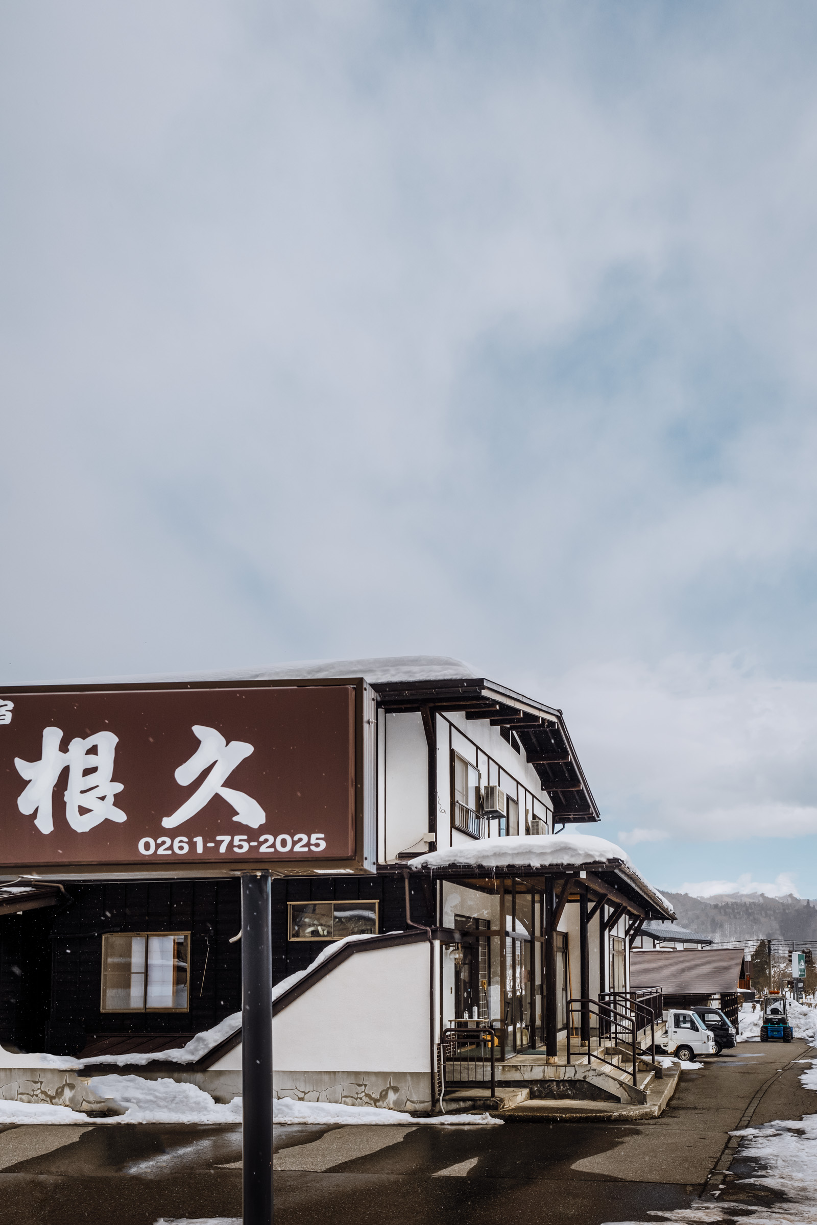 A snow-covered building with a large sign displaying a phone number, set against a cloudy sky. The architecture features traditional Japanese elements.