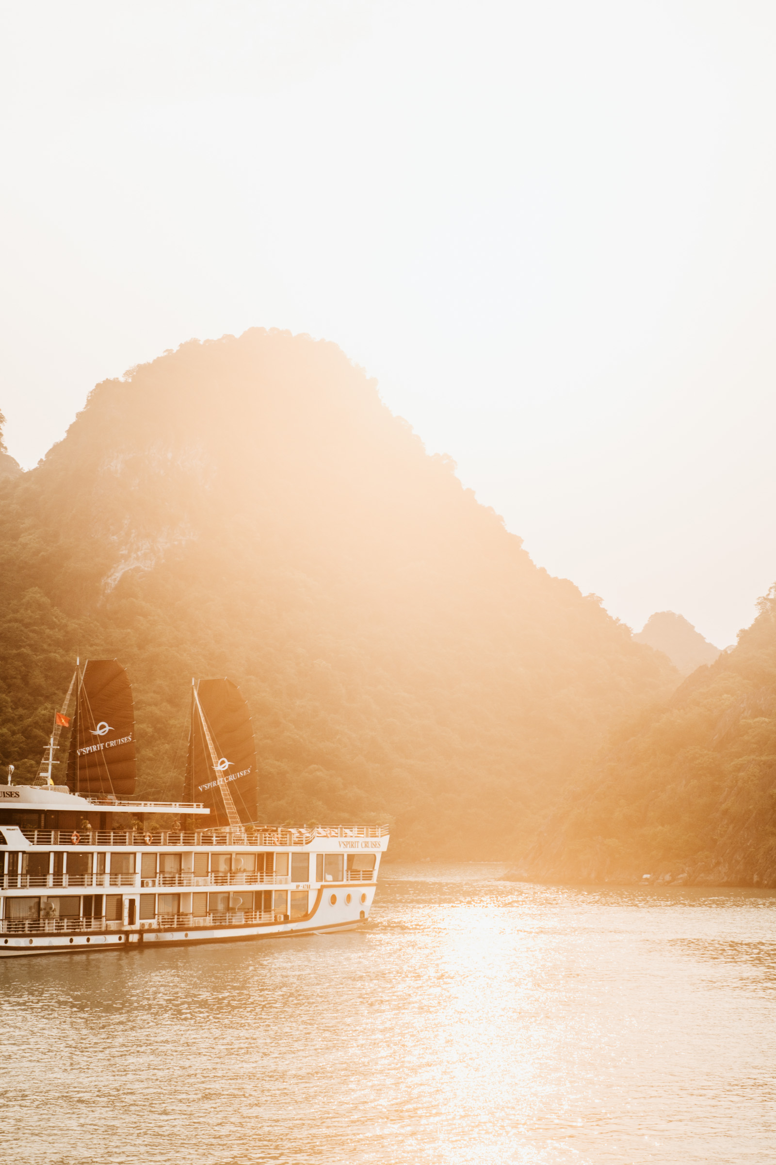 A scenic view of a cruise ship on a tranquil bay, surrounded by misty mountains at sunset, with golden light reflecting on the water.