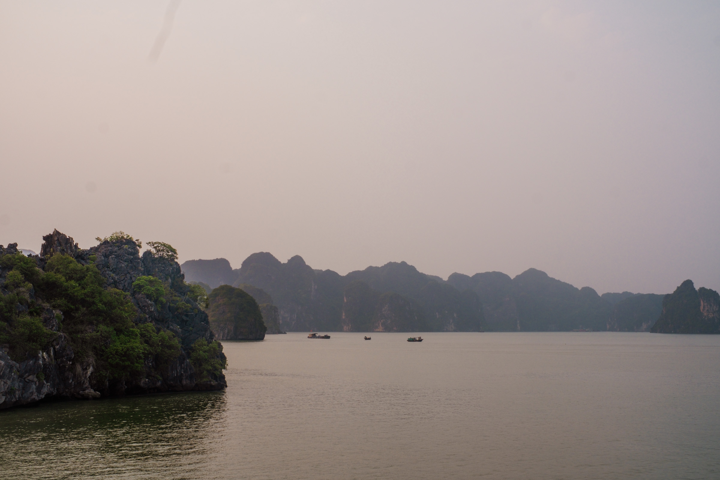 A serene landscape of Ha Long Bay with limestone karsts and boats on calm waters during a hazy sunset.