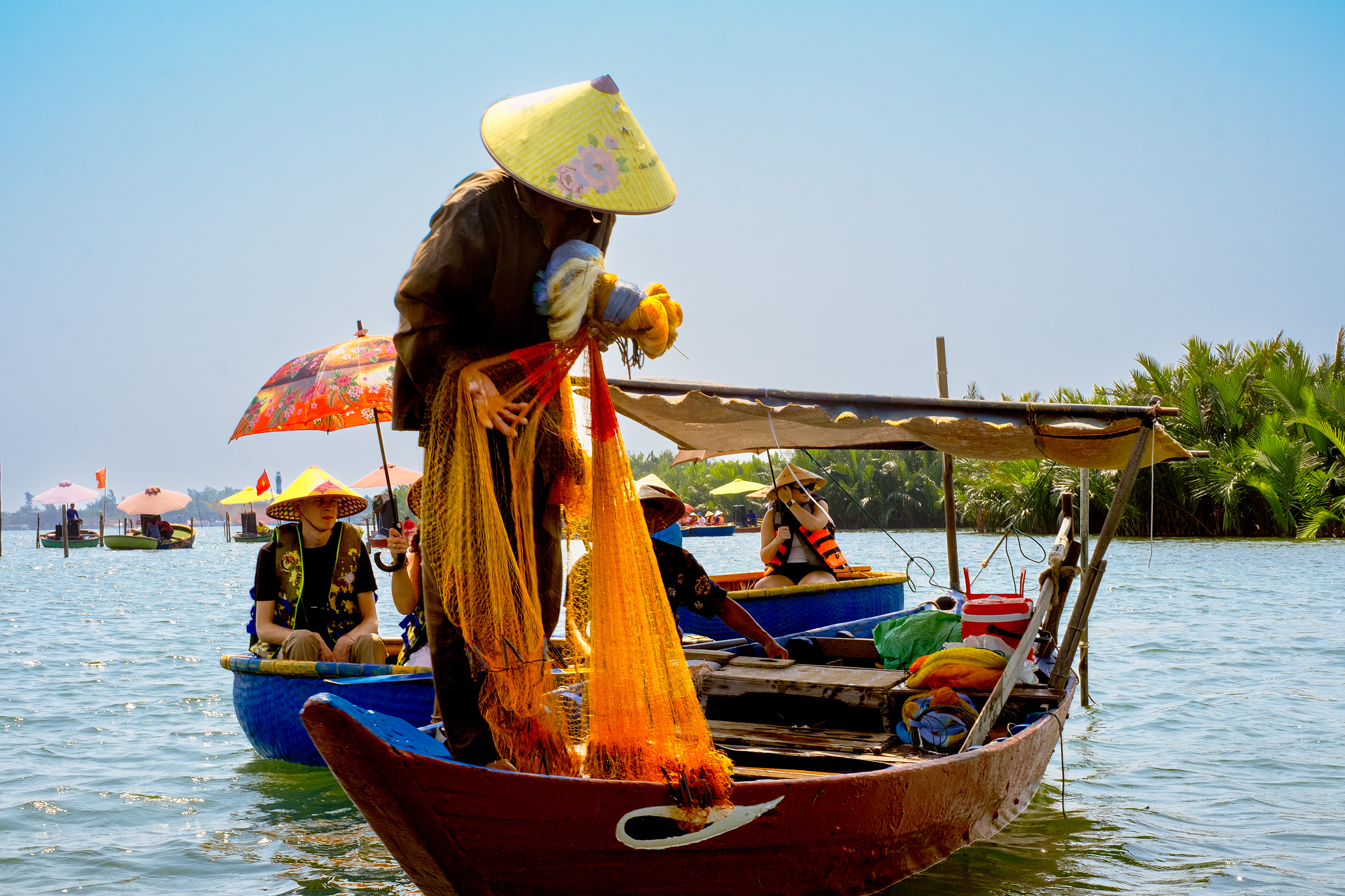 A fisherman in a conical hat pulling a fishing net from a colorful boat on a sunny day, with other fishermen in the background and lush greenery along the water.