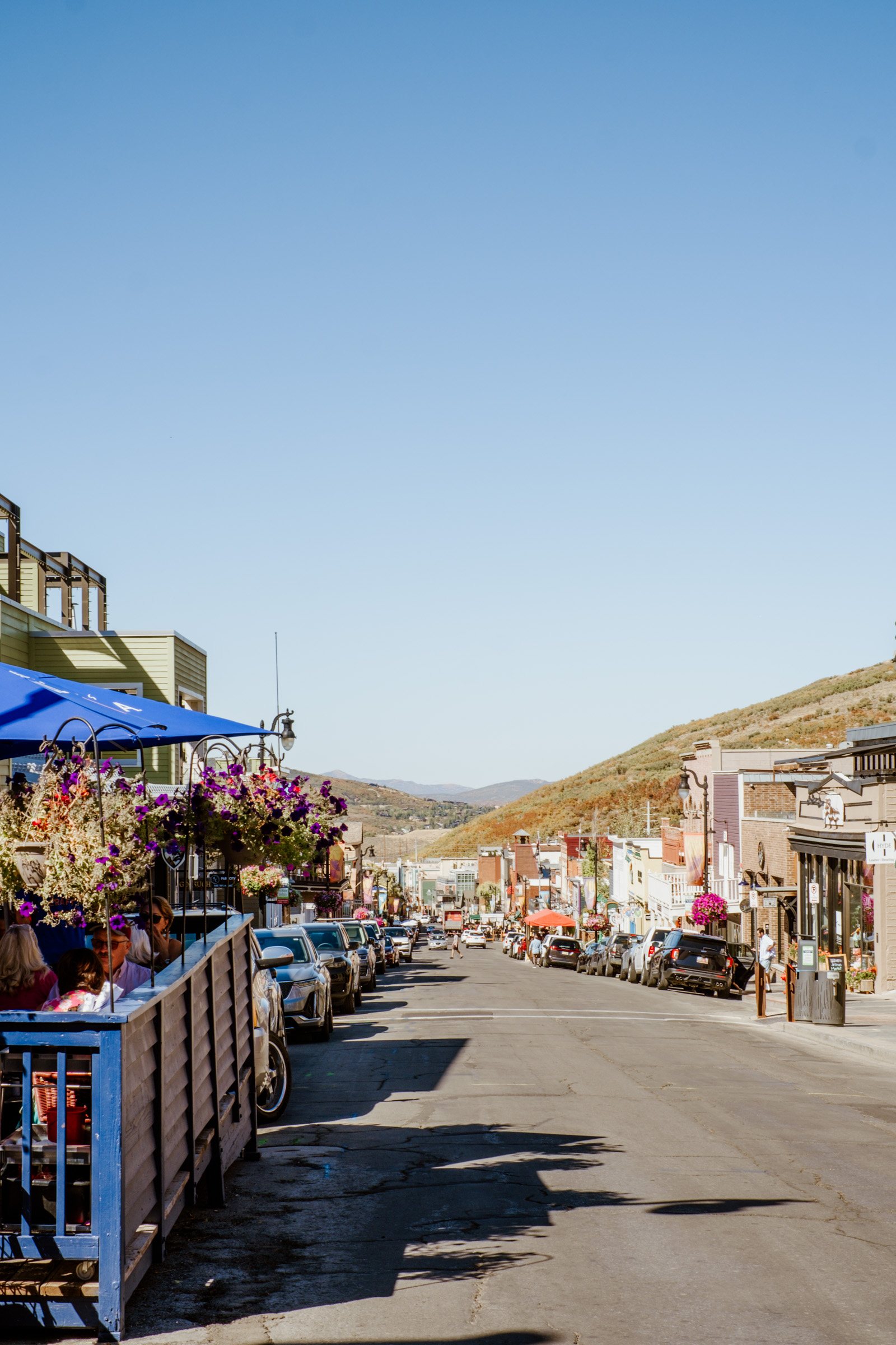A vibrant street scene featuring colorful buildings, parked cars, and outdoor dining areas, with a clear blue sky and hills in the background.