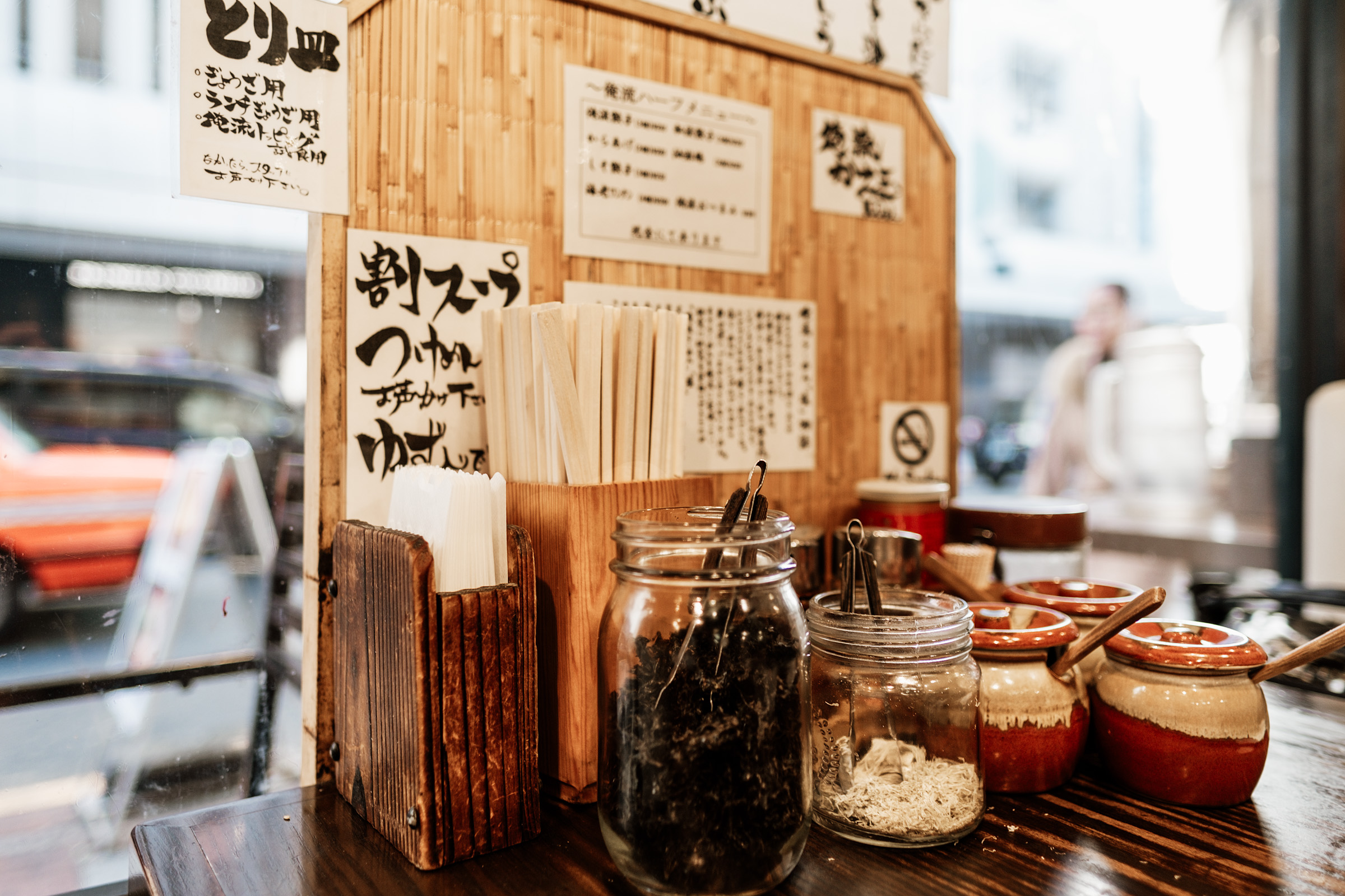 A cozy restaurant interior featuring a wooden counter with jars of condiments and utensils, surrounded by Japanese signage and a view of the street outside.