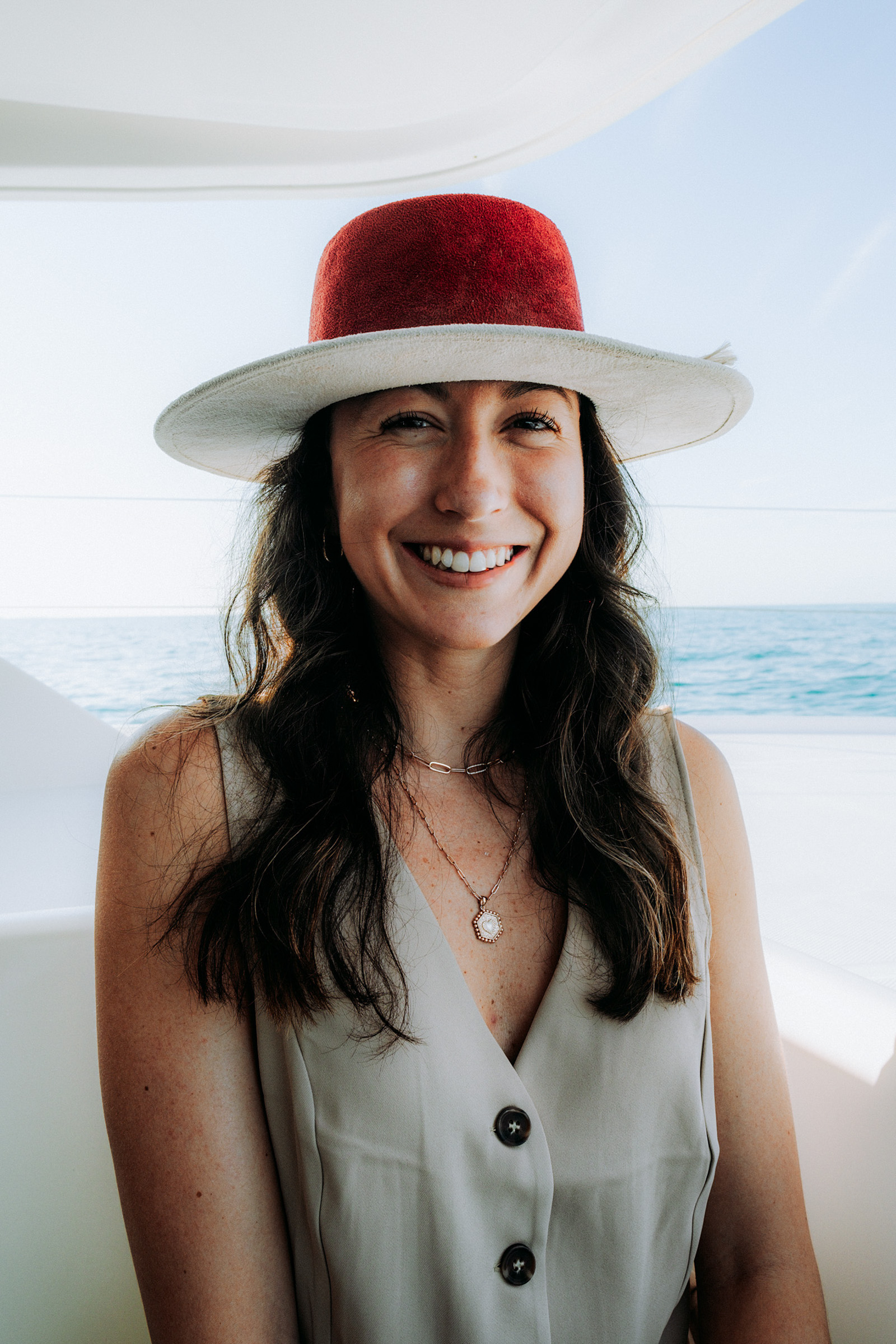 A smiling woman with long brown hair wearing a red hat and a beige sleeveless top, sitting on a boat with the ocean in the background.